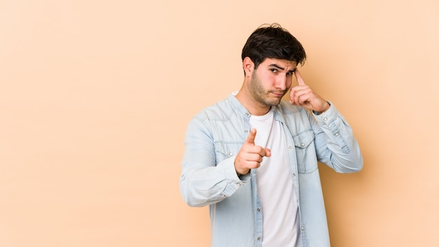 Young man on beige wall pointing temple with finger, thinking