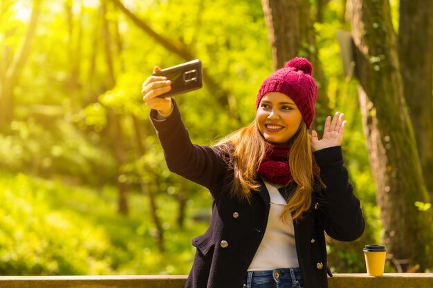 Young man in a black jacket, scarf and red woolen hat enjoying in an autumn park, waving on a video call with the phone