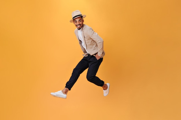 Young man in black pants jacket and hat jumps on orange background Cheerful guy in suit moving on isolated backdrop