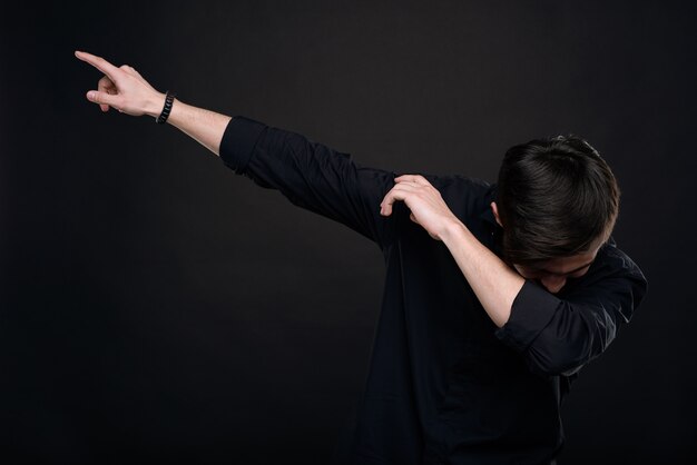 Young man in black shirt dropping head into crook of elbow while making dab move against isolated dark background