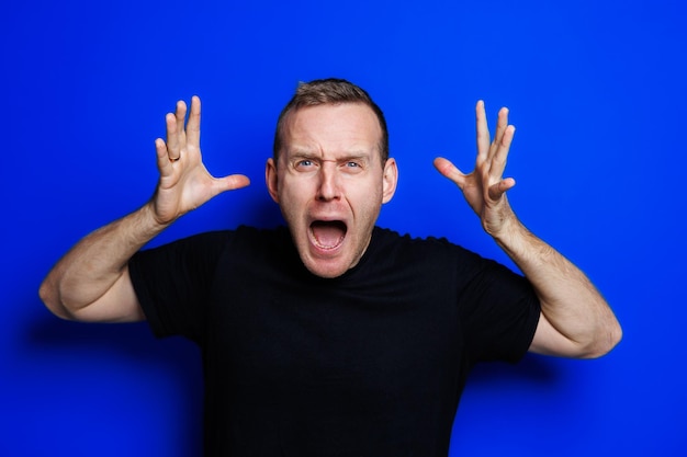 A young man in a black Tshirt on a blue background shows different emotions on his face Cheerful man with a smile Place for text Male beauty