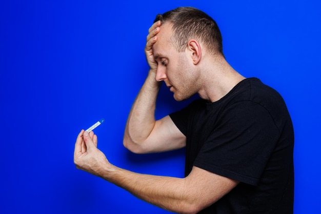 A young man in a black Tshirt on a blue background stands with a thermometer in his hands An increase in body temperature feeling sick Weakened immunity Selective focus