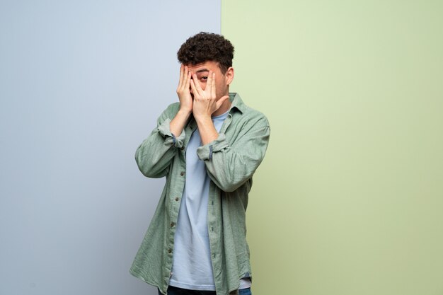 Young man over blue and green  covering eyes by hands and looking through the fingers