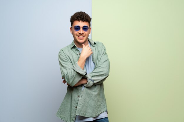 Young man over blue and green wall with glasses and smiling