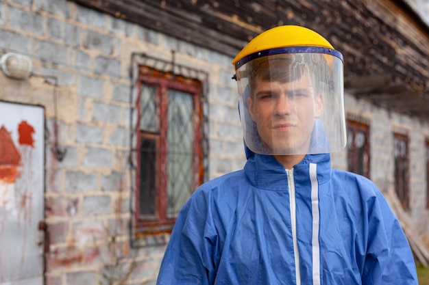 青い防護服とプラスチック製医療用マスクの若い男が建物の近くに立っています。