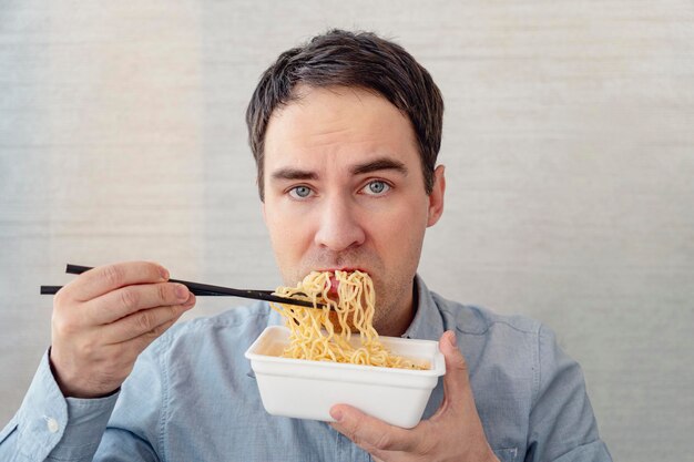 青いシャツを着た若い男が不満そうな顔で箱から麺を食べている オフィスでの昼食 味のないジャンク フード