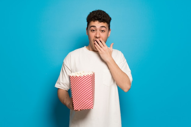 Young man over blue  surprised and eating popcorns