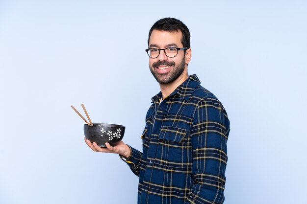 Young man over blue wall smiling a lot while holding a bowl of noodles with chopsticks