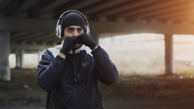 Young man boxer in headphones training punches in urban location outdoors in winter