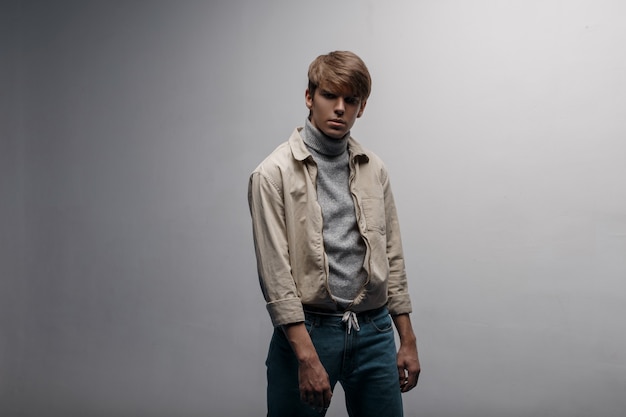 Young man in a bright light stylish jacket in a knitted gray golf in blue trendy pants in retro style is standing in a room near the white wall.European stylish guy. Spring modern men's fashion