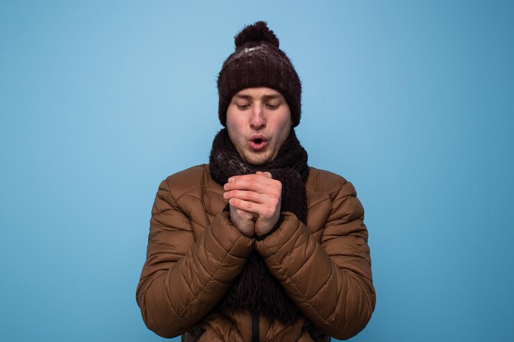 A young man in a brown scarf and winter hat, a light brown jacket on a blue background warms his hands