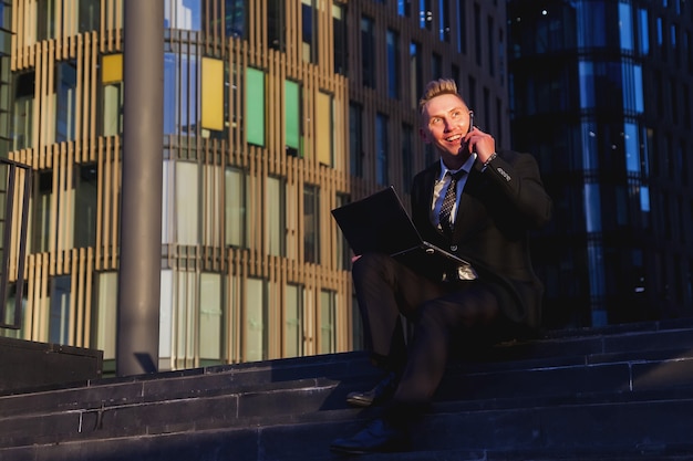 Young man businessman in style wear clothes, suit sitting on steps with laptop