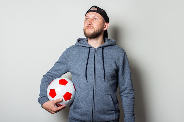 Young man in a cap and hoodie holds a soccer ball and looks to the side against a light wall.