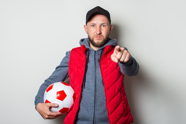 Young man in a cap, hoodie and vest holds a soccer ball and points his finger at the viewer