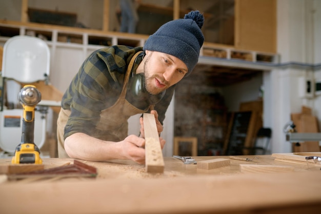 Young man carpenter blowing sawdust from wooden plank. Woodworking at modern carpentry workshop