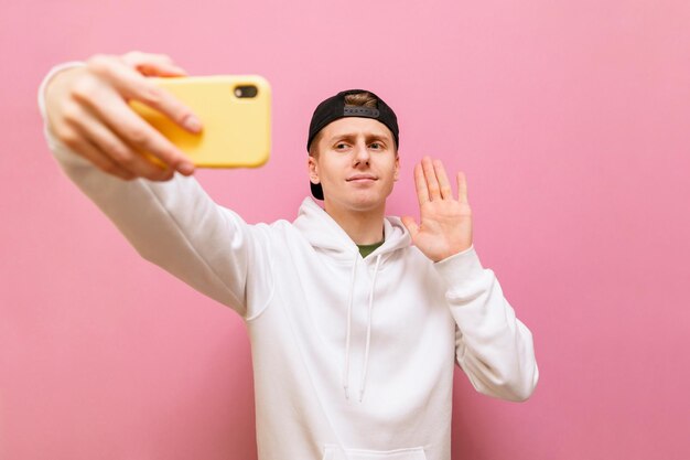 Young man in casual clothes makes a video call on a smartphone on a pink background looks in camera of phone and is greeted with a palm raised Guy makes a phone call and shows quotHelloquot gesture