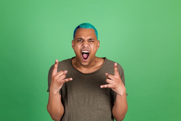 Young man in casual on green wall blue hair happy screaming with rock gesture