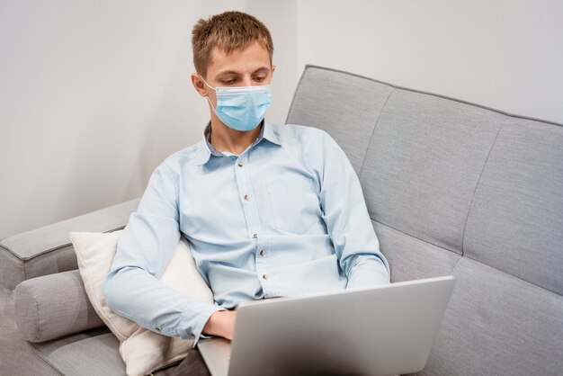 A young man of caucasian appearance in a blue shirt and a protective mask with a laptop on the couch...