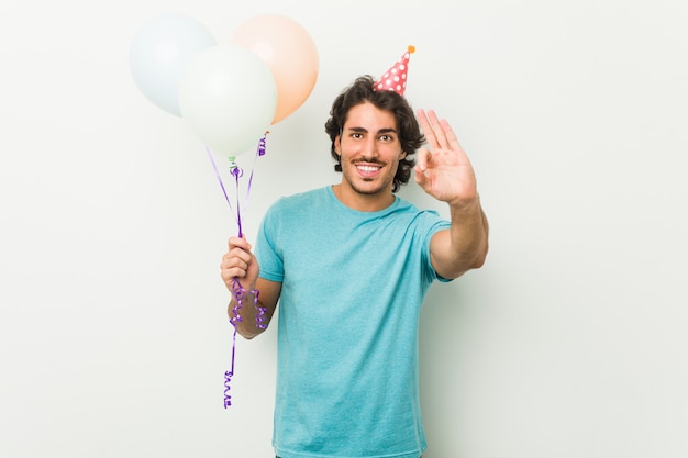 Young man celebrating a party holding balloons cheerful and confident showing ok gesture.