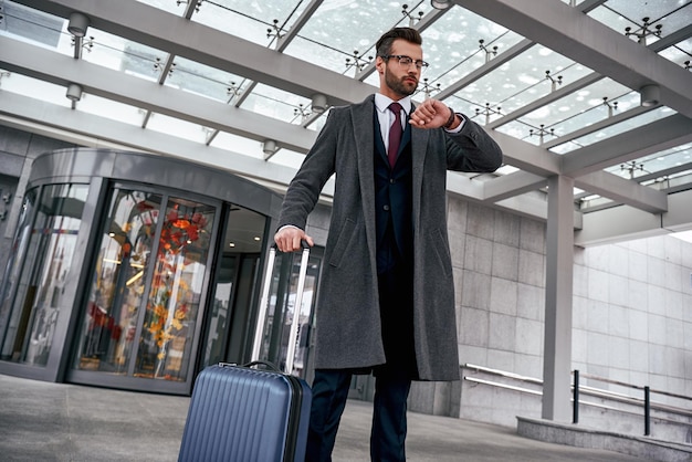 Young man checking his watch and pulling suitcase