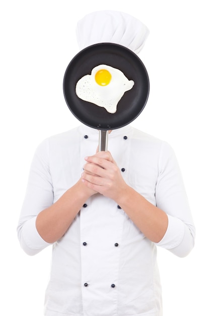 Young man in chef uniform holding skillet with frying egg behind her face isolated on white background