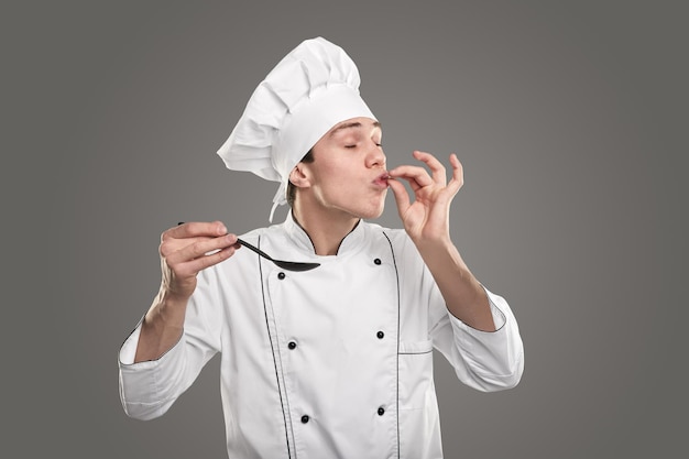 Young man in chef uniform kissing fingers with closed eyes after tasting delicious food in spoon against gray background