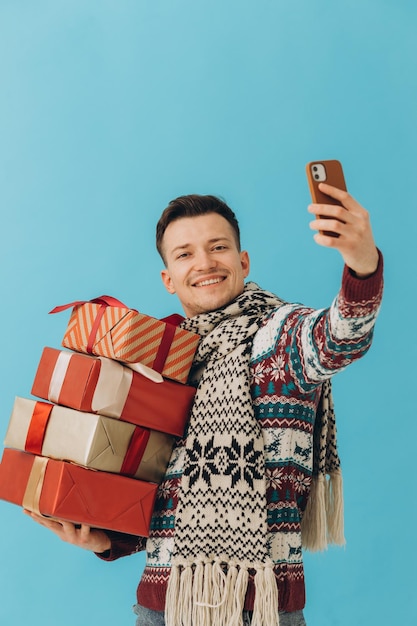 Young man in Christmas sweater and scarf holding many gift boxes and taking selfie isolated on blue background Happy new year celebration concept