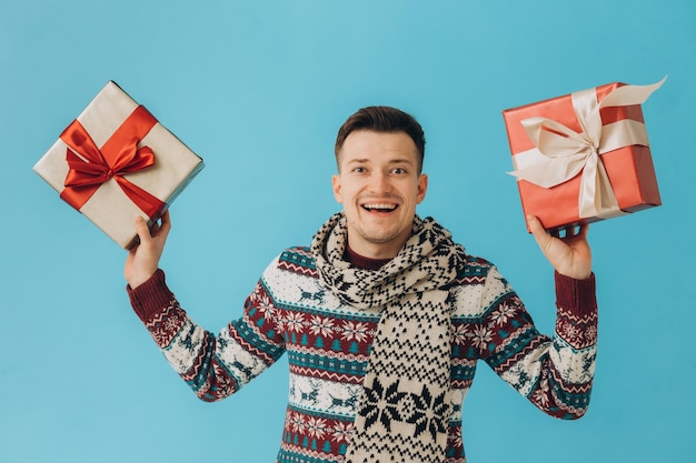 Young man in christmas sweater and scarf holding many gift\
boxes with gift ribbon bow isolated on blue background happy new\
year celebration concept