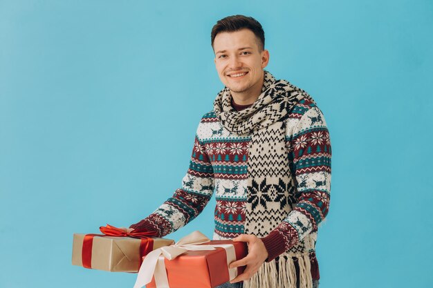 クリスマス セーターとスカーフ ギフト リボン付きの多くのギフト ボックスを保持している若い男は、青の背景に分離された弓を弓します。新年あけましておめでとうございますお祝いコンセプト