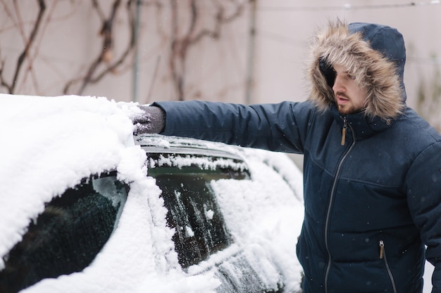 Young man cleaning snow from car windshield outdoors near the garage on winter day