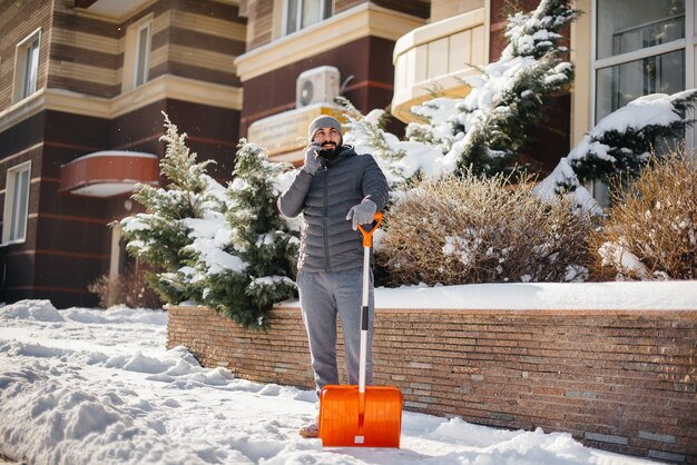 Молодой человек расчищает снег перед домом в солнечный и морозный день и разговаривает по телефону.