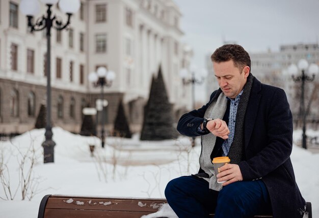 A young man in a coat and suit sits on a bench in a city park looks at his watch with a cup of coffee winter season