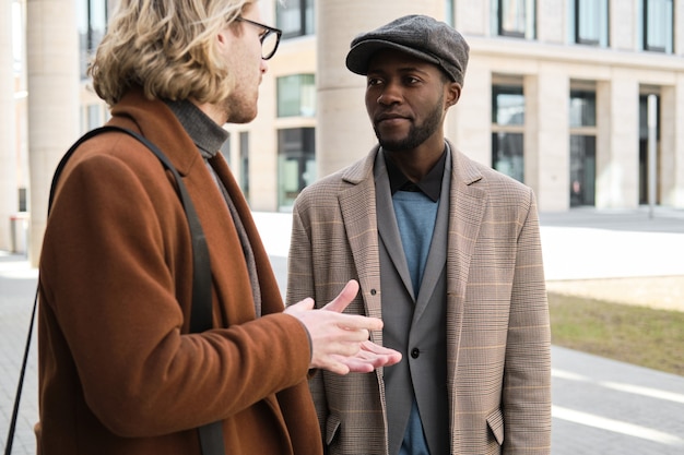 Young man in coat talking to his colleague while they standing in the city