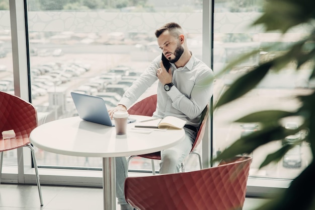 若い男は、カフェで携帯電話で話し、彼のラップトップに集中して取り組んでいます。