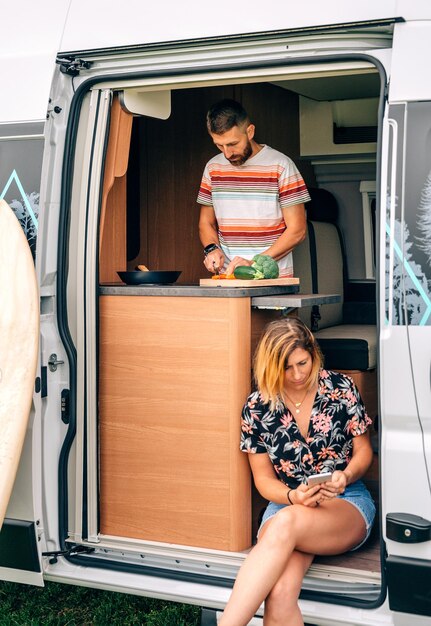 Photo young man cooking in a camper van while his wife looks at the mobile