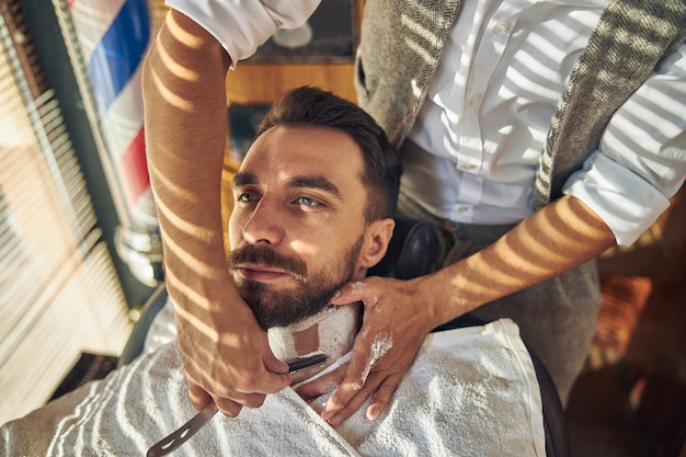 Foto un giovane ricoperto di crema che si fa la barba con un rasoio a mano libera da un barbiere professionista