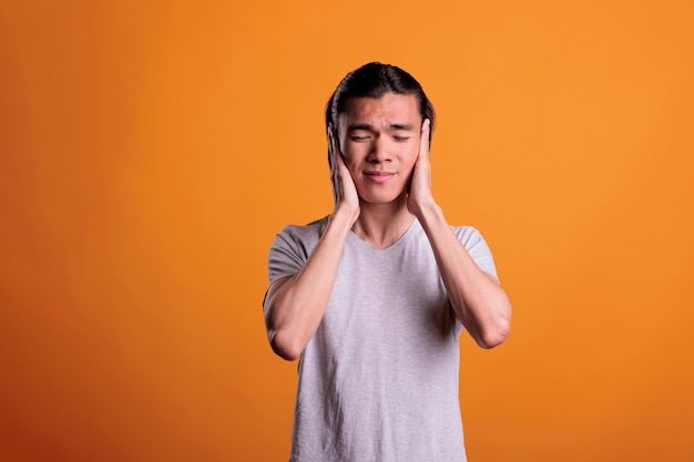 Young man covering ears with hands, hear no evil, three wise\
monkeys moral concept. asian teenager with closed eyes showing no\
sound gesture, front view studio medium shot