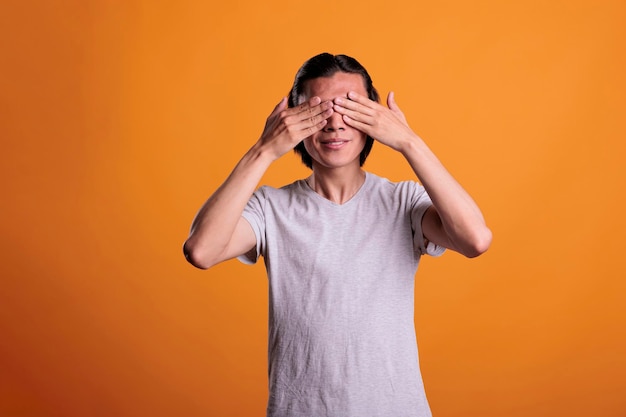 Young man covering eyes with hands, see no evil, three wise monkeys concept, blindness sign. Smiling asian teenager standing, showing no look gesture front view medium shot