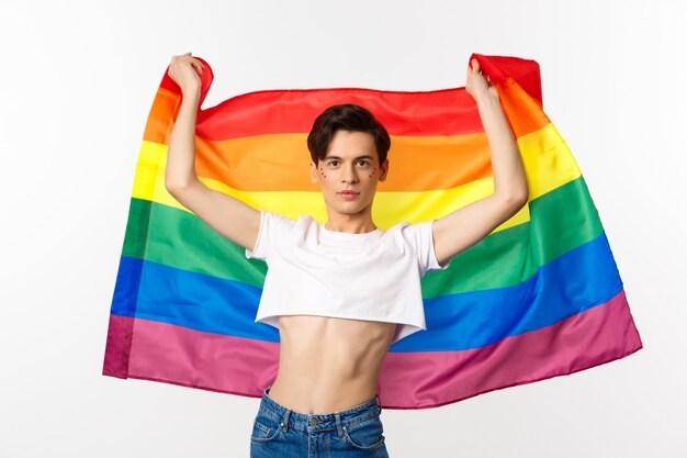 Young man in crop top, with glitter on face, raising pride flag with confident emotion. Queer person smiling with a lgbt flag