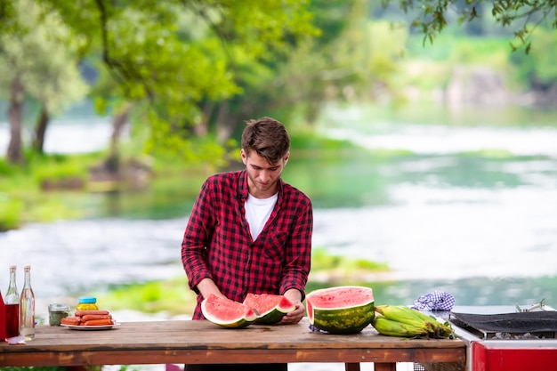 young man cutting juicy watermelon during outdoor french dinner party near the river on beautiful summer evening in the nature