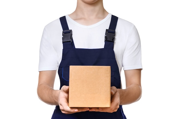 Young man in dark blue overalls and and a white T-shirt holding out cardboard box in front of him, isolated on white wall. Delivery concept.