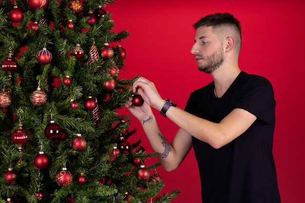 Young man decorating xmas tree on red background. Christmas New Year's Eve preparation, holiday stress relief planning tips, winter season tradition, single celebration, alone concept