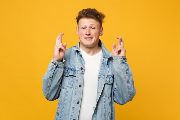 Young man in denim casual clothes waiting for special moment, keeping fingers crossed, making wish isolated on yellow orange background. people sincere emotions lifestyle concept. mock up copy space