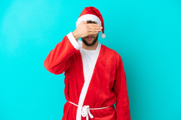 Young man disguised as Santa Claus isolated on blue background covering eyes by hands. Do not want to see something