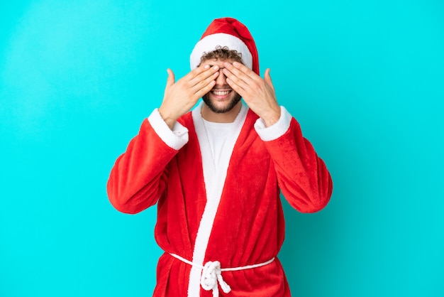 Young man disguised as Santa Claus isolated on blue background covering eyes by hands