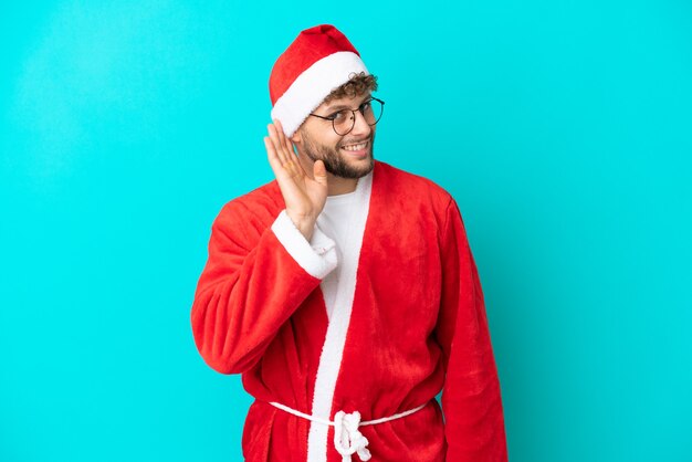 Young man disguised as Santa Claus isolated on blue background listening to something by putting hand on the ear