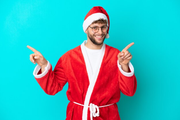 Giovane travestito da babbo natale isolato su sfondo blu che punta il dito verso i laterali e felice