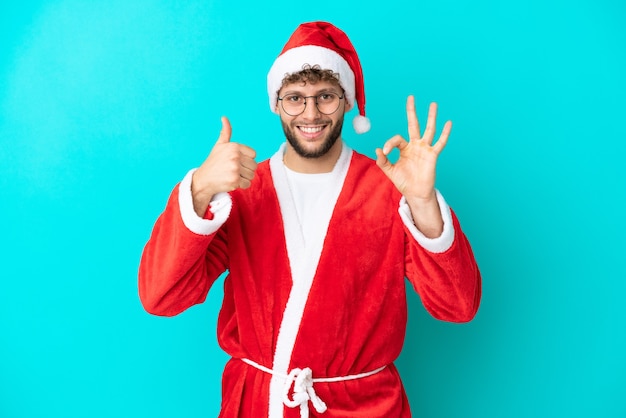 Young man disguised as Santa Claus isolated on blue background showing ok sign and thumb up gesture