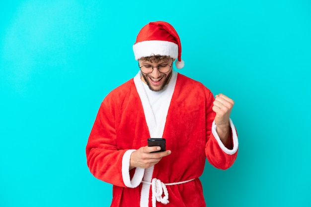 Young man disguised as Santa Claus isolated on blue background surprised and sending a message