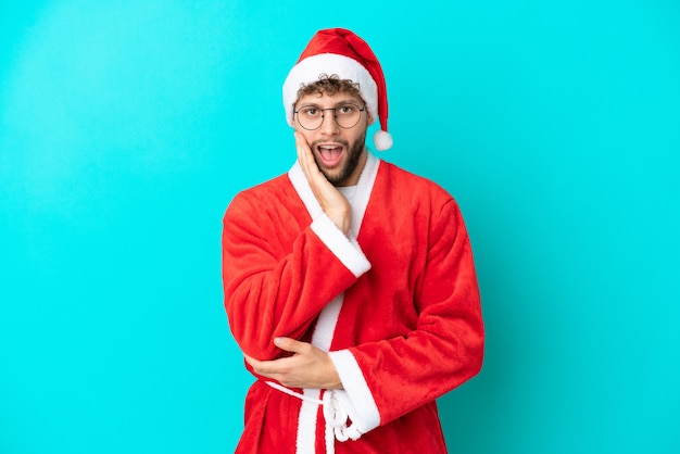 Young man disguised as Santa Claus isolated on blue background surprised and shocked while looking right